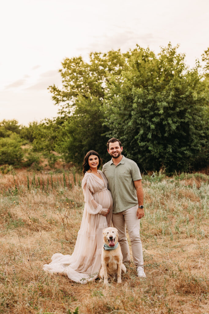 An expecting wife and husband standing in a field at sunset with their golden retriever. 