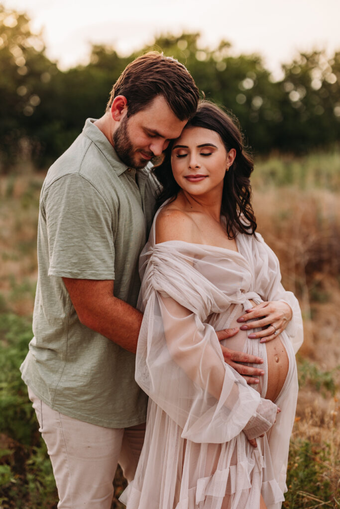 Husband and wife standing in a field at sunset holding the wife's pregnant belly. 