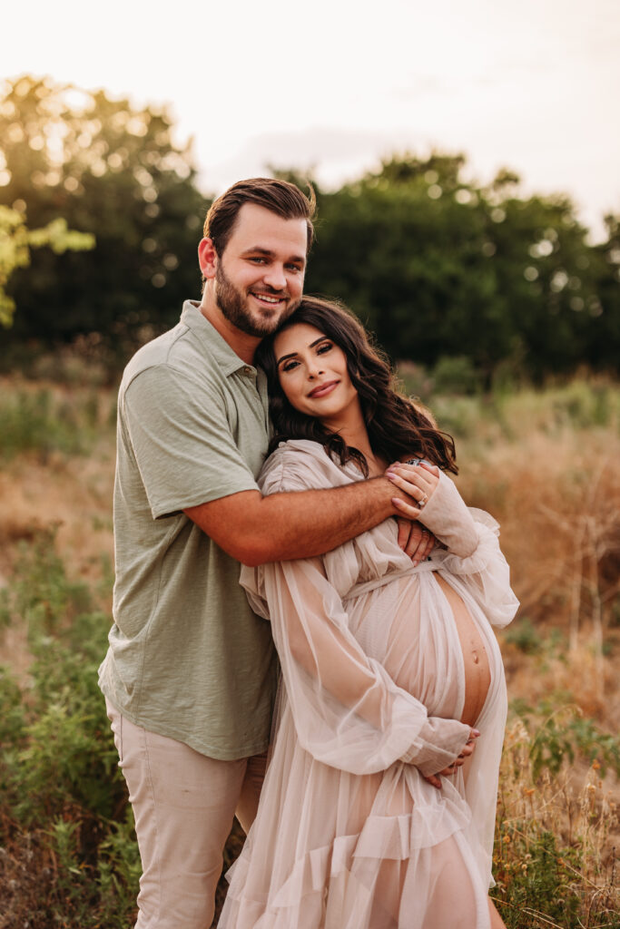 A husband and wife standing together while the wife holds her pregnant belly. 