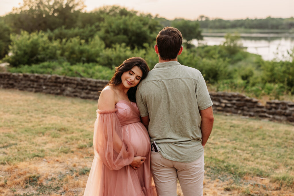 Expecting mother holds her belly and leans on her husbands shoulder at golden hour. 