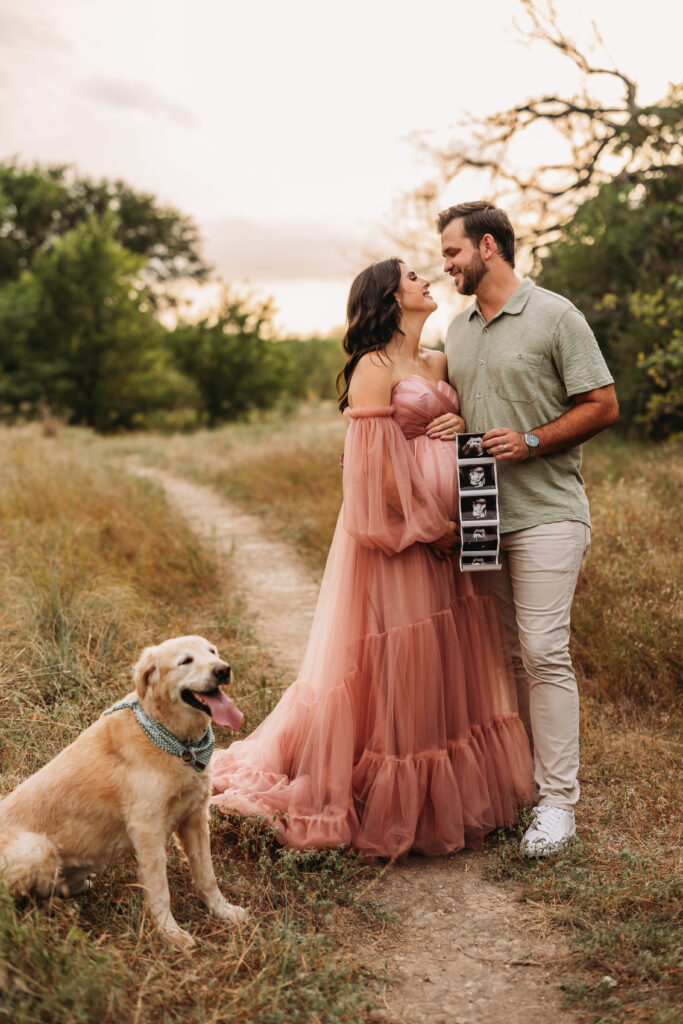 Parents hold up a sonogram photo while their golden retriever sits close by. 